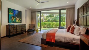 Un dormitorio con cama, ventilador de techo, ventana grande, un cuadro, cómoda y una silla.