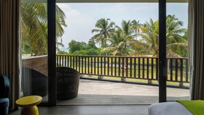 Habitación de hotel con balcón, una cama, una mesa amarilla y vista a palmeras y un cuerpo de agua.