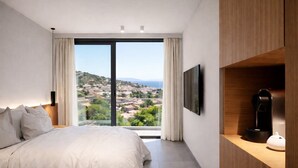 Ein Schlafzimmer mit einem Bett, einem Fernseher, einer Kaffeemaschine und Blick auf eine Stadt durch große Fenster.