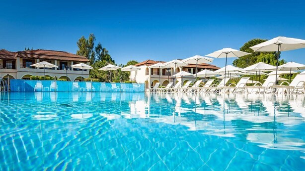 Ein klares Schwimmbecken mit Liegestühlen und Sonnenschirmen, ein Gebäude mit Ziegeldach und Grünpflanzen im Hintergrund.