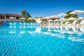 En pool med klart vatten, där det finns solstolar och parasoller, en byggnad med ett tak täckt av tegelpannor och grönska i bakgrunden.