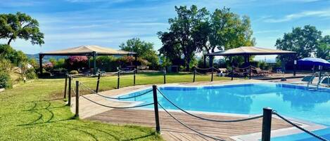 A swimming pool with a wooden deck, surrounded by trees and covered seating areas.