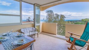 Ein Balkon mit Esstisch für vier Personen, einem Stuhl und Blick auf das Meer und B äume.
