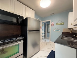 A kitchen with a stainless steel refrigerator, a microwave, and an oven. There are white cabinets and a blue wall with a 'BEACH HOUSE' sign.