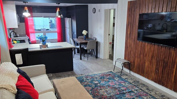 Sala de estar con sofá, reposapiés y una alfombra grande. Zona de cocina con refrigerador, fregadero y mesa de comedor. Un televisor está montado en la pared.