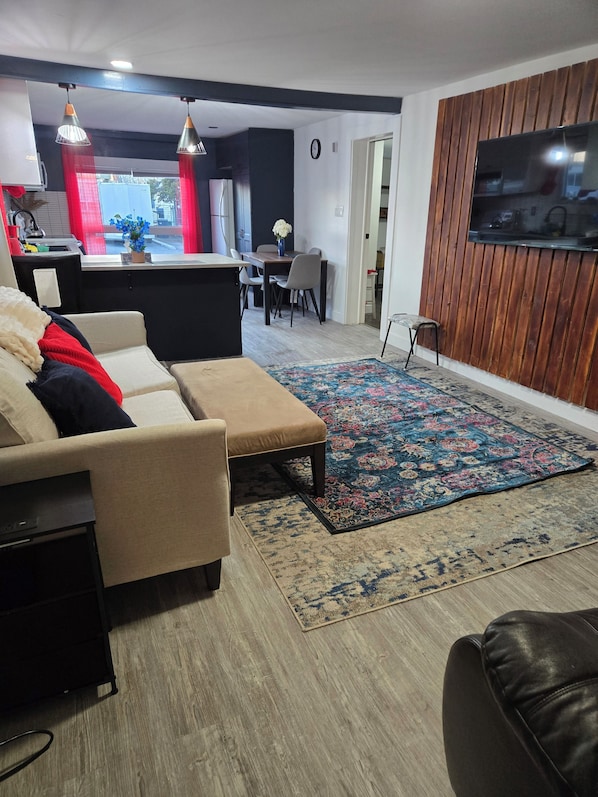Una sala de estar con un sofá, una mesa de centro, un tapete, un televisor y una cocina al fondo.