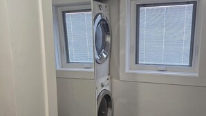 Un lavadero con lavadora y secadora de carga frontal, una ventana con persianas y piso de madera oscura.