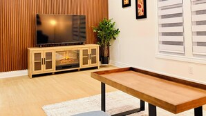 Un salon avec un téléviseur, un mur recouvert de panneaux de bois, deux tableaux encadrés, une plante et une fenêtre avec des stores.