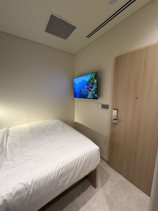 Une chambre à coucher avec un lit, un téléviseur fixé au mur, une porte en bois et une bouche d’aération au plafond.