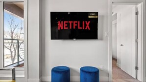 Habitació amb un televisor de pantalla plana gran fixat a la paret que mostra el logotip de Netflix, dos tamborets blaus tapissats i una porta que condueix a una altra habitació.
