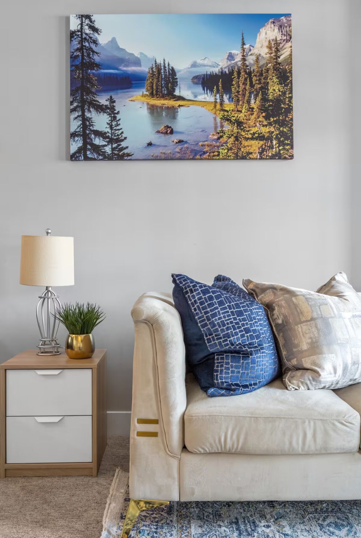 Une pièce avec un canapé beige, une petite table de chevet en bois sur laquelle se trouvent une lampe et une plante, et un grand tableau représentant un paysage accroché au mur.