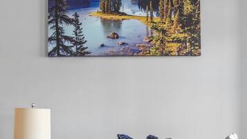 Une pièce avec un canapé beige, une petite table de chevet en bois sur laquelle se trouvent une lampe et une plante, et un grand tableau représentant un paysage accroché au mur.