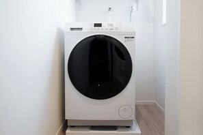Un appareil combiné lave-linge et sèche-linge à chargement frontal, de couleur blanche, doté d’un panneau de commande, placé dans un coin, avec un plancher en bois et des murs blancs.
