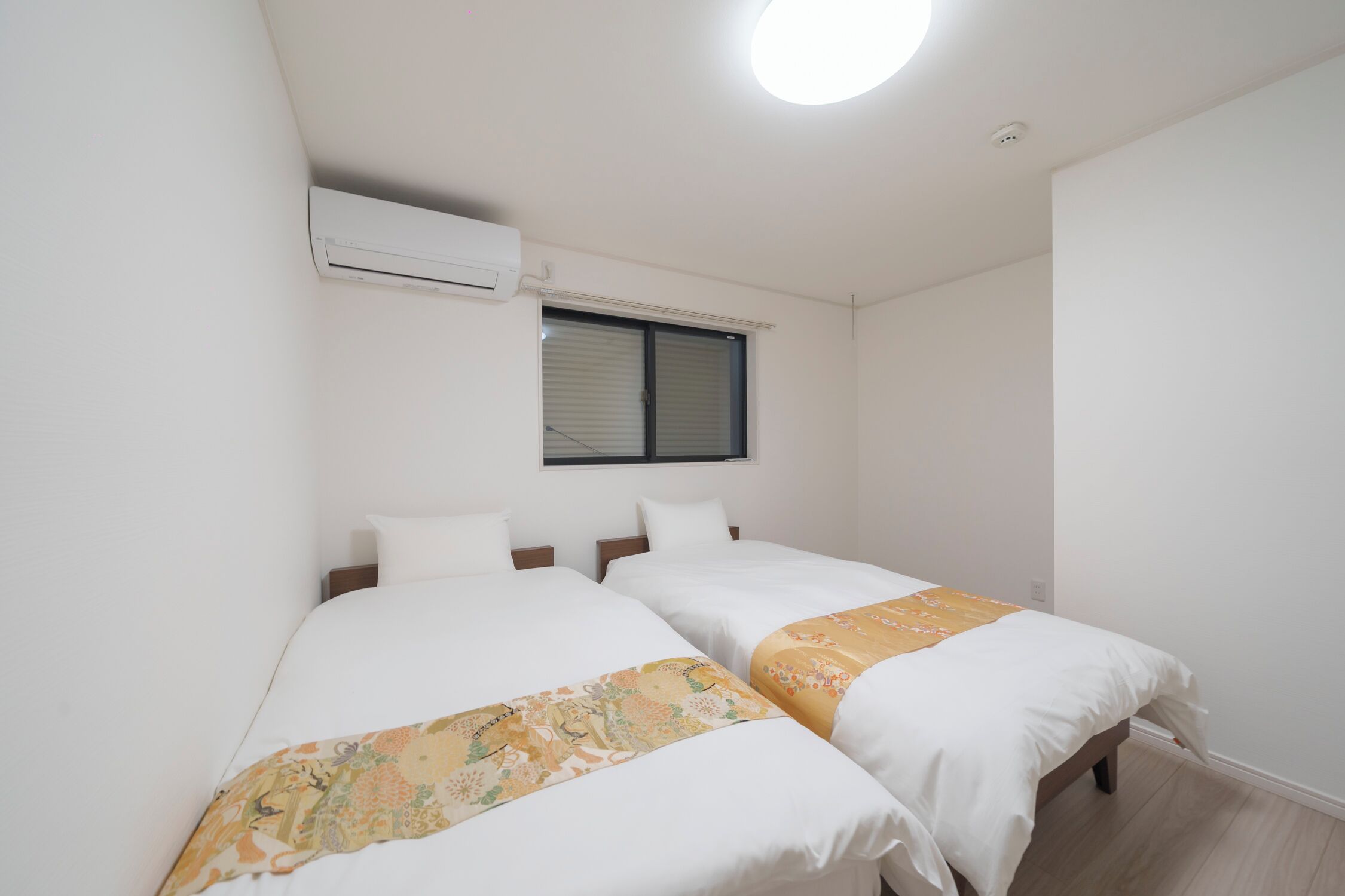 A twin room with two beds, a window with blinds, an air conditioning unit, and a ceiling light.