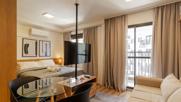 A modern hotel room with a bed, a TV, a desk with a chair, a sofa, and large windows with curtains. - Studio Climatizado a 100m da Esta o Pinheiros (São Paulo)