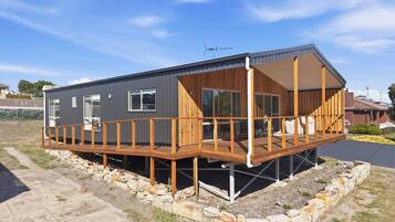 Una cabaña moderna de dos pisos con terraza de madera, ventanas grandes y techo de metal.