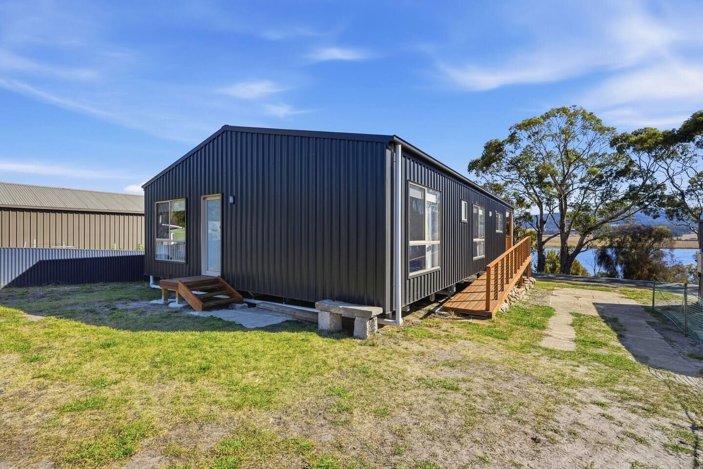 A compact, dark-colored cabin with a wooden deck, a small outdoor seating area, and a view of a body of water.
