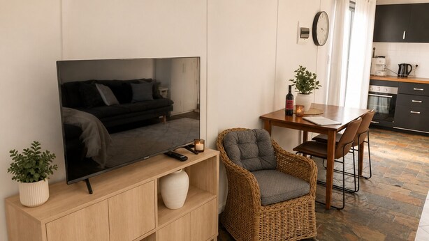 Un salon moderne avec un téléviseur à écran plat, un fauteuil en osier, une table à manger en bois et une cuisine en arrière-plan.