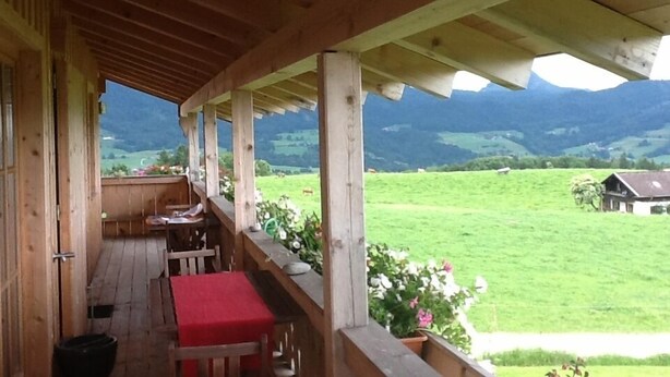 Une véranda en bois avec une table et des chaises rouges, donnant sur un paysage verdoyant avec des collines et une maison.