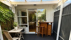 A covered patio with a wooden deck, a table with chairs, and a small bar area. - Coastal Retreat with Private Verandah -
King Bed - Walk to Beach & Train (Adelaide)