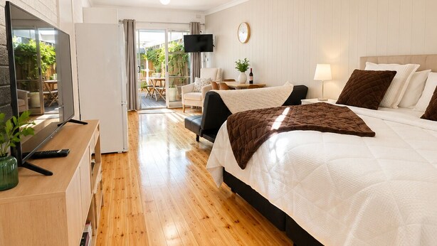 A modern hotel room with a wooden floor, a large bed, a desk with a flat-screen TV, and a view of the outdoors. - Coastal Retreat with Private Verandah -
King Bed - Walk to Beach & Train (Adelaide)