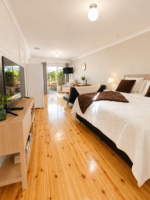 A modern hotel room with a wooden floor, a large bed, a desk with a flat-screen TV, and a view of the outdoors. - Coastal Retreat with Private Verandah -
King Bed - Walk to Beach & Train (Adelaide)