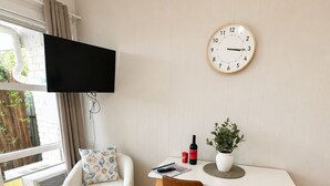 A cozy room with a white sofa, a dining table with two chairs, a wall-mounted clock, and a TV on the wall. - Coastal Retreat with Private Verandah -
King Bed - Walk to Beach & Train (Adelaide)