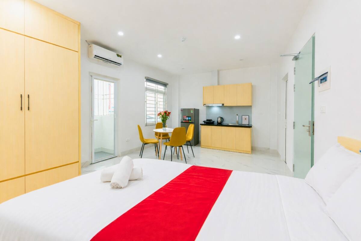 A hotel room with a bed, a red runner, a dining table with chairs, a kitchenette, and a wardrobe.