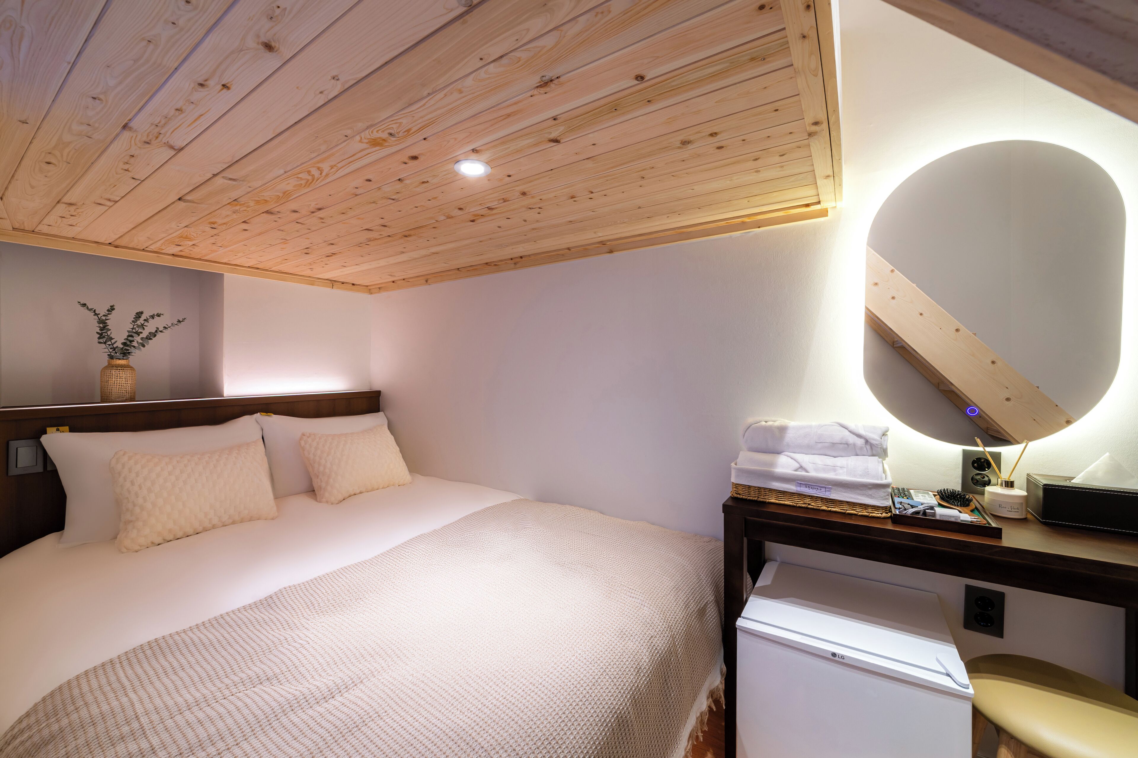Un dormitorio con techo de madera, una cama con cubrecama blanco, una mesita de noche con lámpara y un pequeño electrodoméstico blanco.