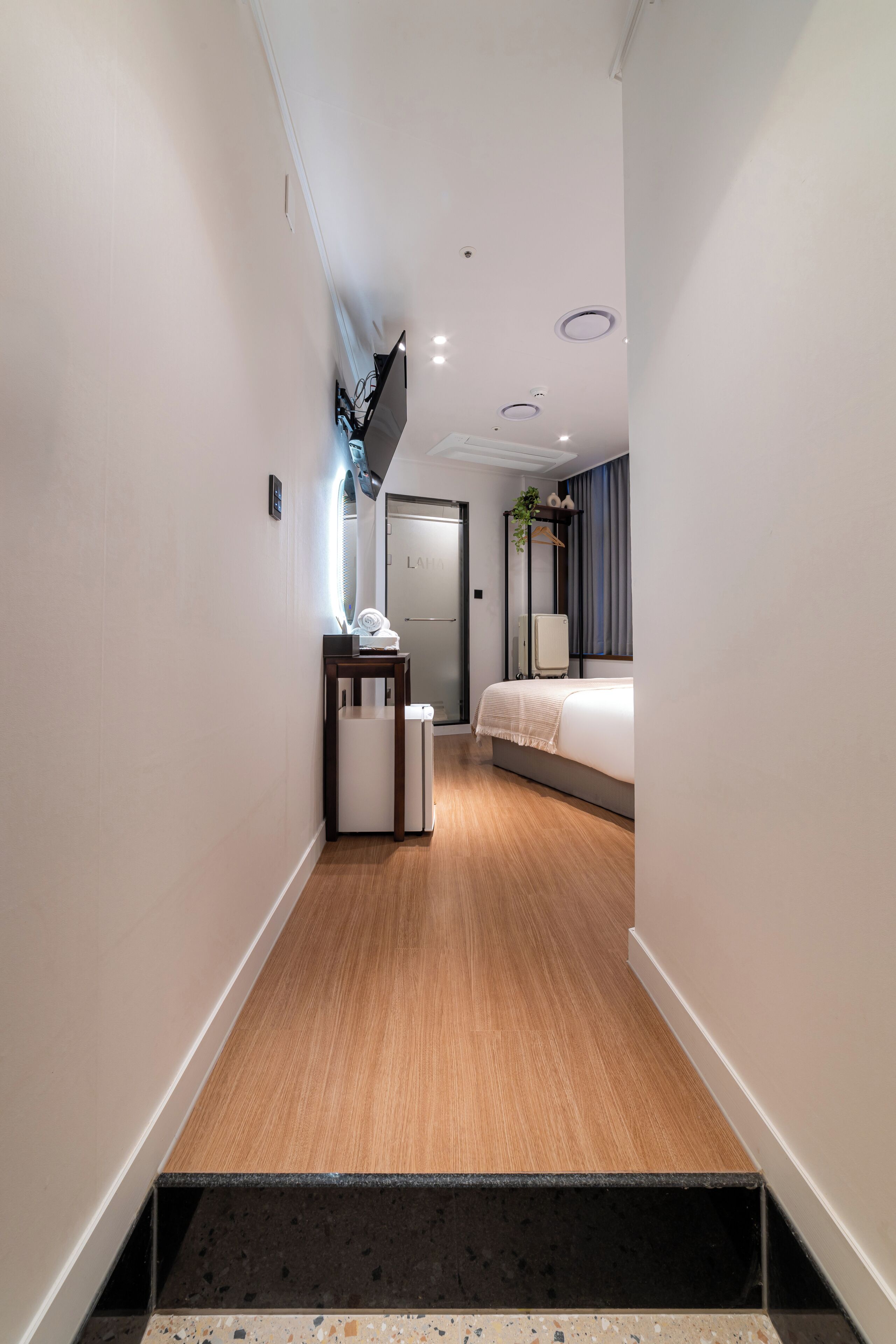 Um corredor estreito com piso de madeira, uma pequena mesa com abajur, uma porta e uma cama com mesa de cabeceira ao fundo.