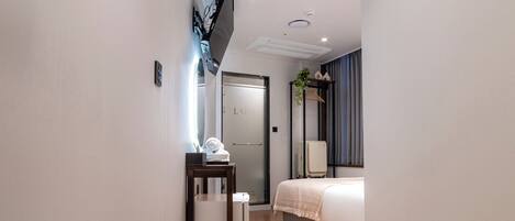 Um corredor estreito com piso de madeira, uma pequena mesa com abajur, uma porta e uma cama com mesa de cabeceira ao fundo.