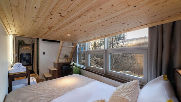 Une chambre avec un plafond en bois, un grand lit, un bureau avec une chaise et une fenêtre donnant sur des arbres et des immeubles.