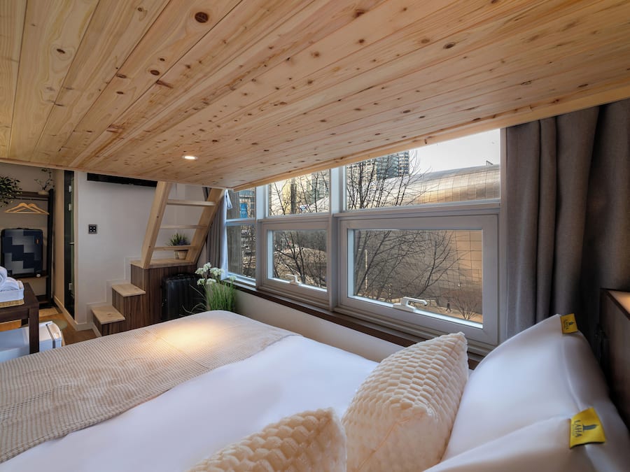 Un dormitorio con techo de madera, una cama grande, un escritorio con silla y una ventana con vista a árboles y edificios.