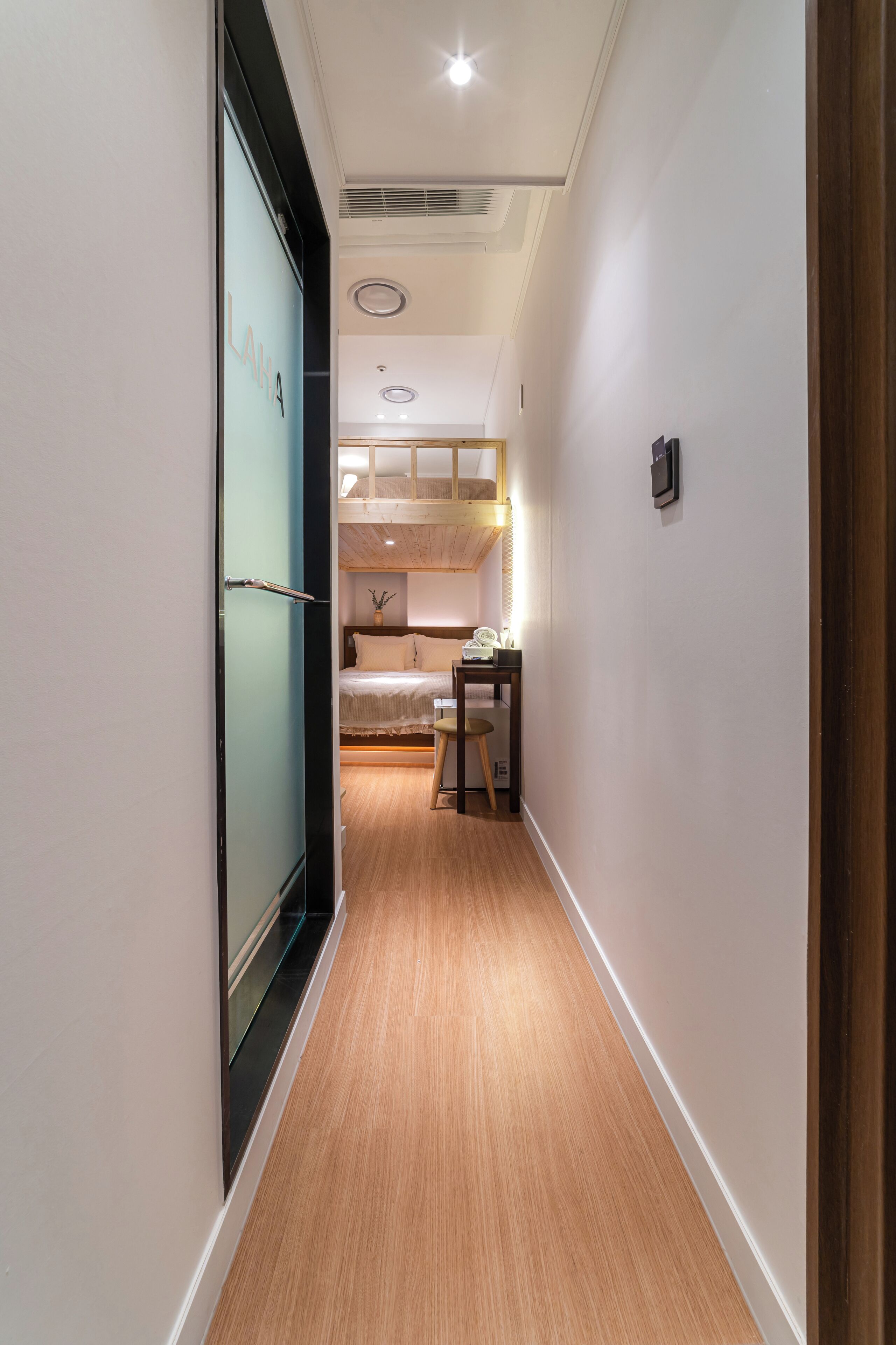 A narrow hallway with a wooden floor, a bed with a white headboard, a desk with a chair, and a small table with a plant.