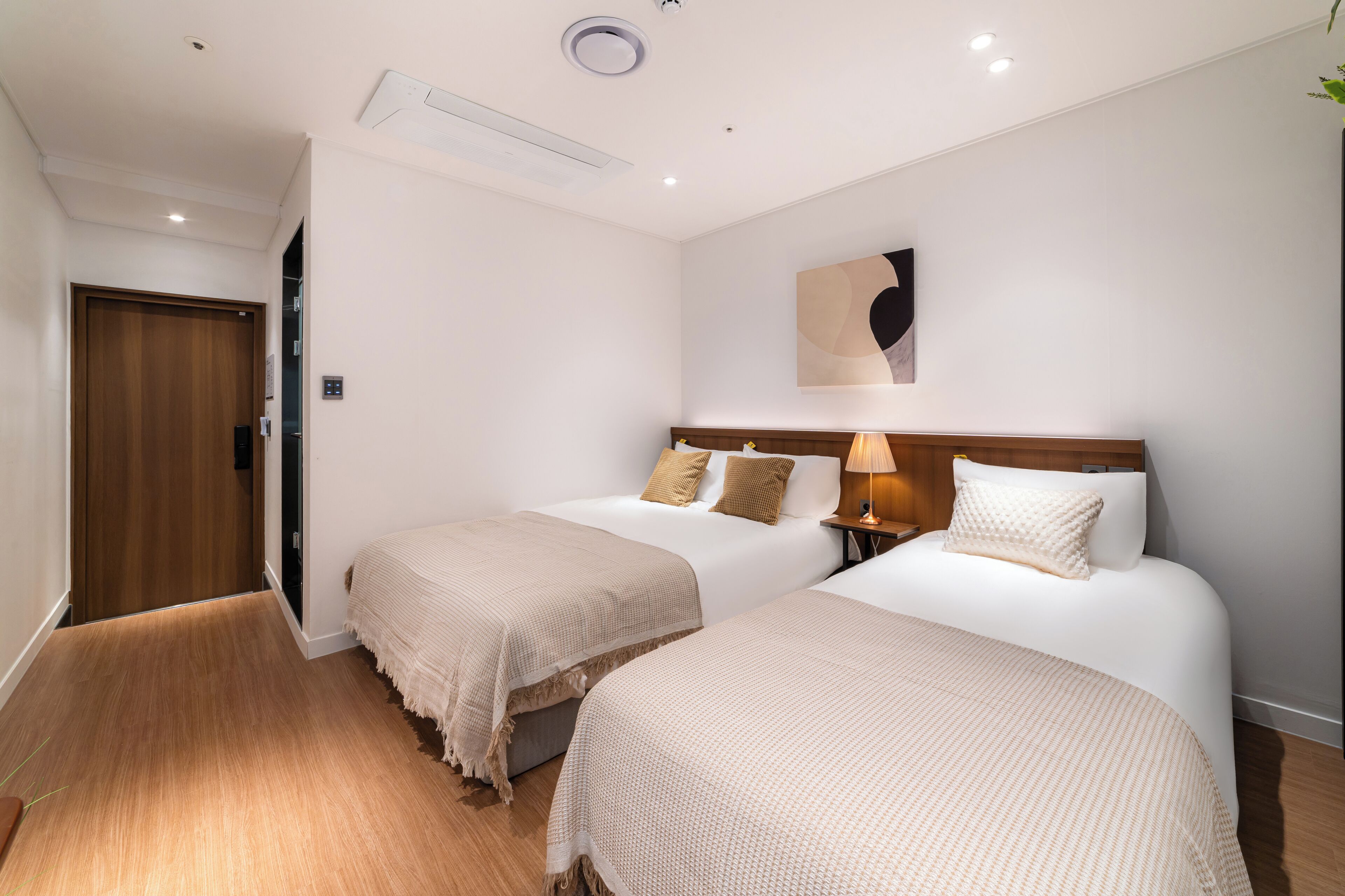 A hotel room with two beds, a wooden door, a wall-mounted air conditioner, and a bedside lamp.