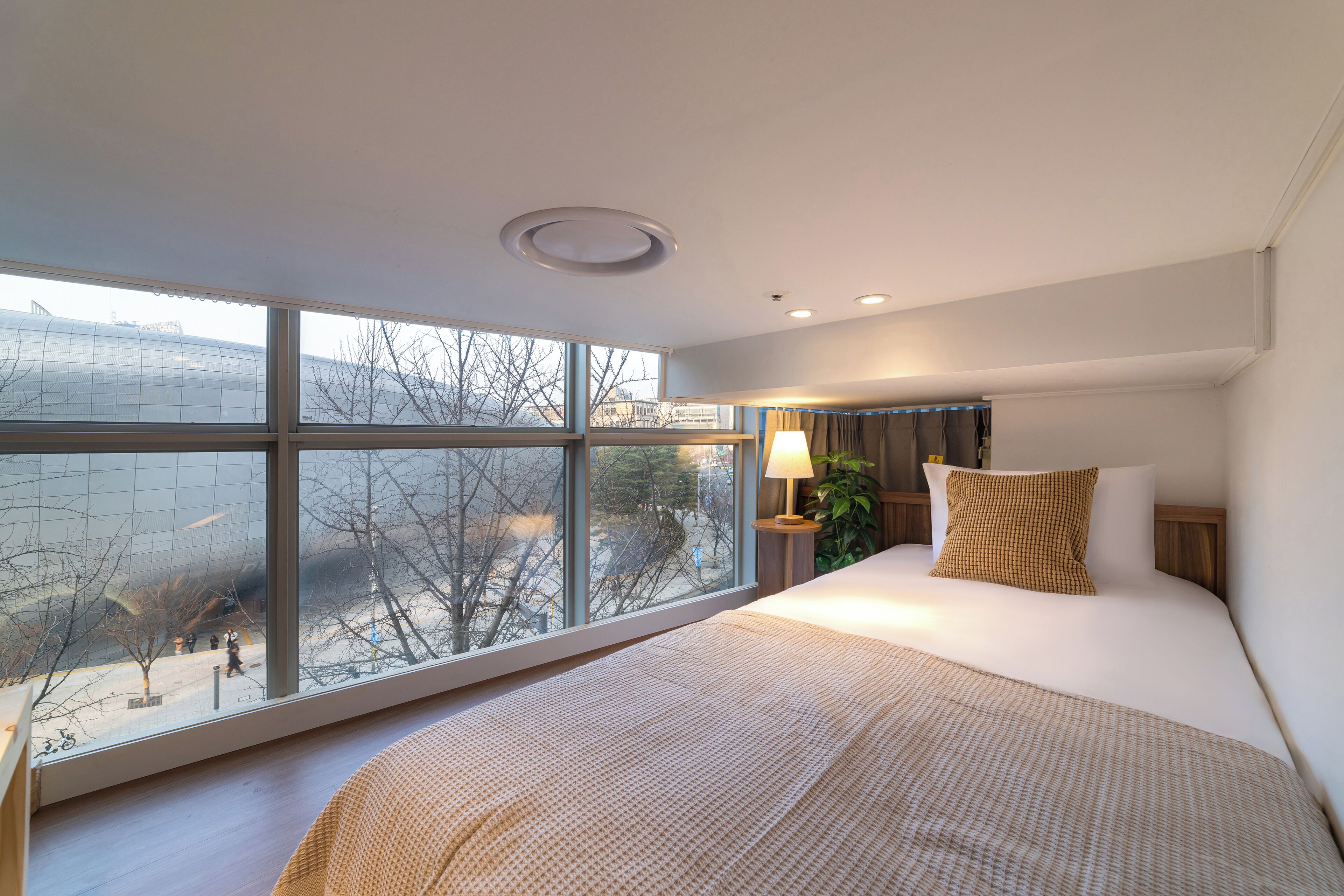 Quarto de hotel moderno com uma janela grande, uma cama com colcha estampada, um abajur e vista para o exterior.