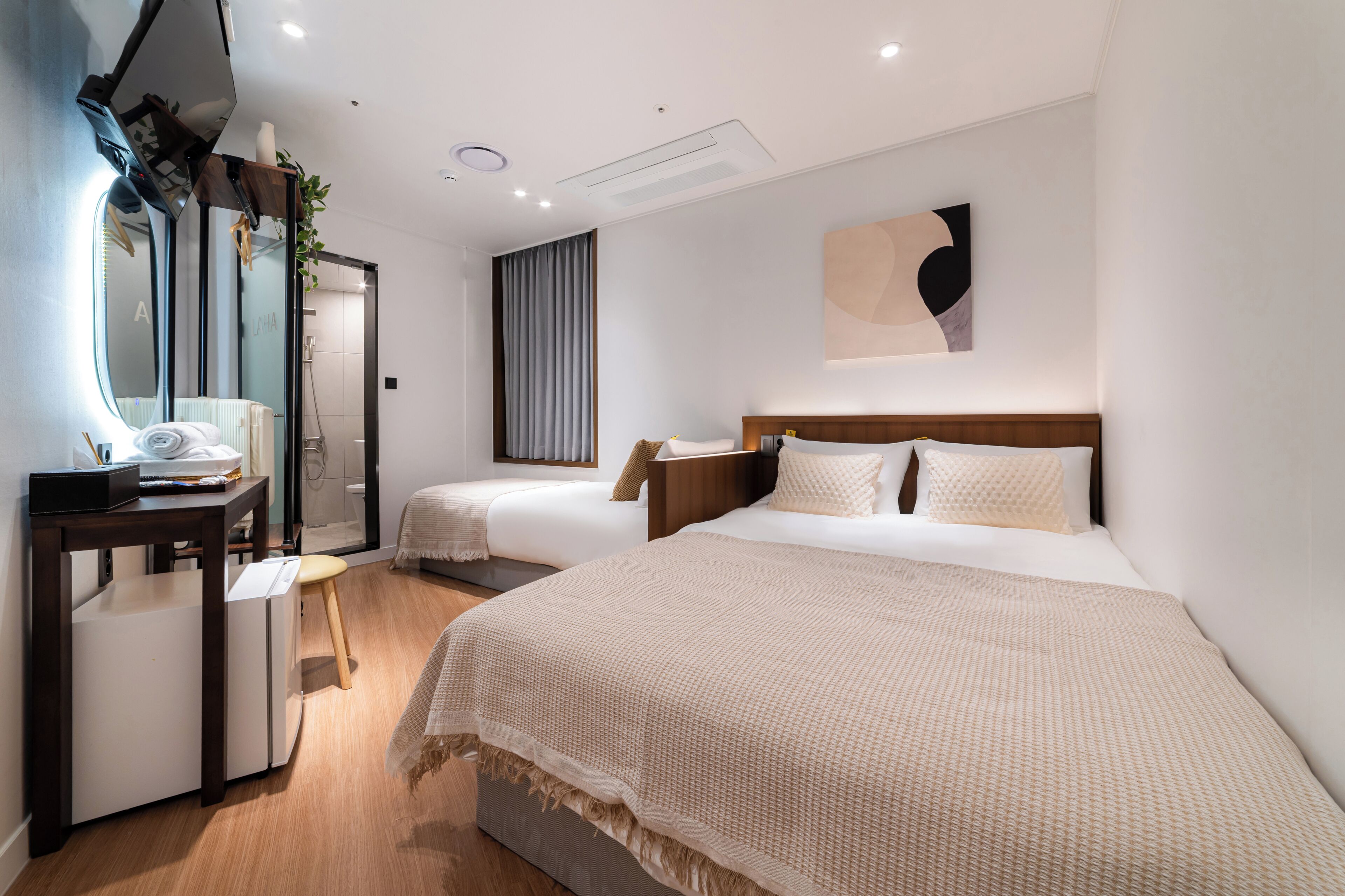 A hotel room with a large bed, a smaller bed, a desk with a chair, a mirror, and a wall-mounted artwork.