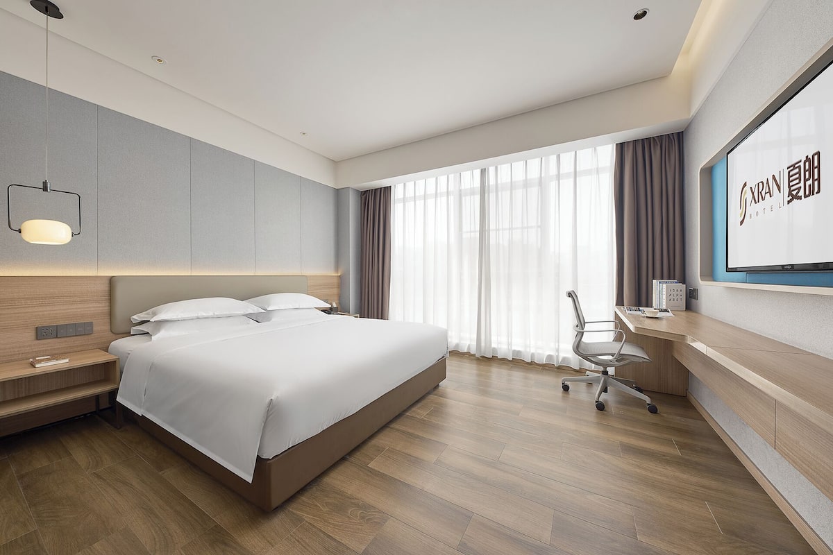A modern hotel room with a bed, bedside table, desk, chair, and large windows.
