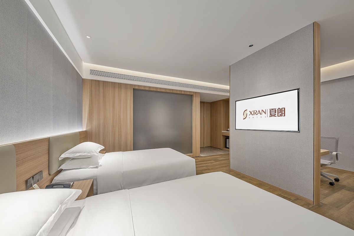 A hotel room with a bed, bedside tables, a desk, a chair, and a wall-mounted TV displaying the hotel name.