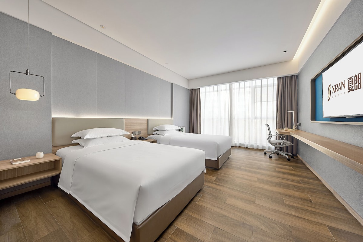 A hotel room with two beds, a desk, a chair, a TV, and a large window with curtains.