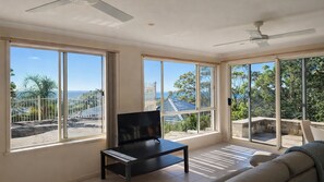 Amplia habitación con grandes ventanas que ofrecen vistas al océano, un ventilador de techo y un mueble negro para televisor.