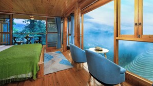 Una cabaña de madera con una cama, sillas, una mesita y ventanas amplias que dan al bosque.