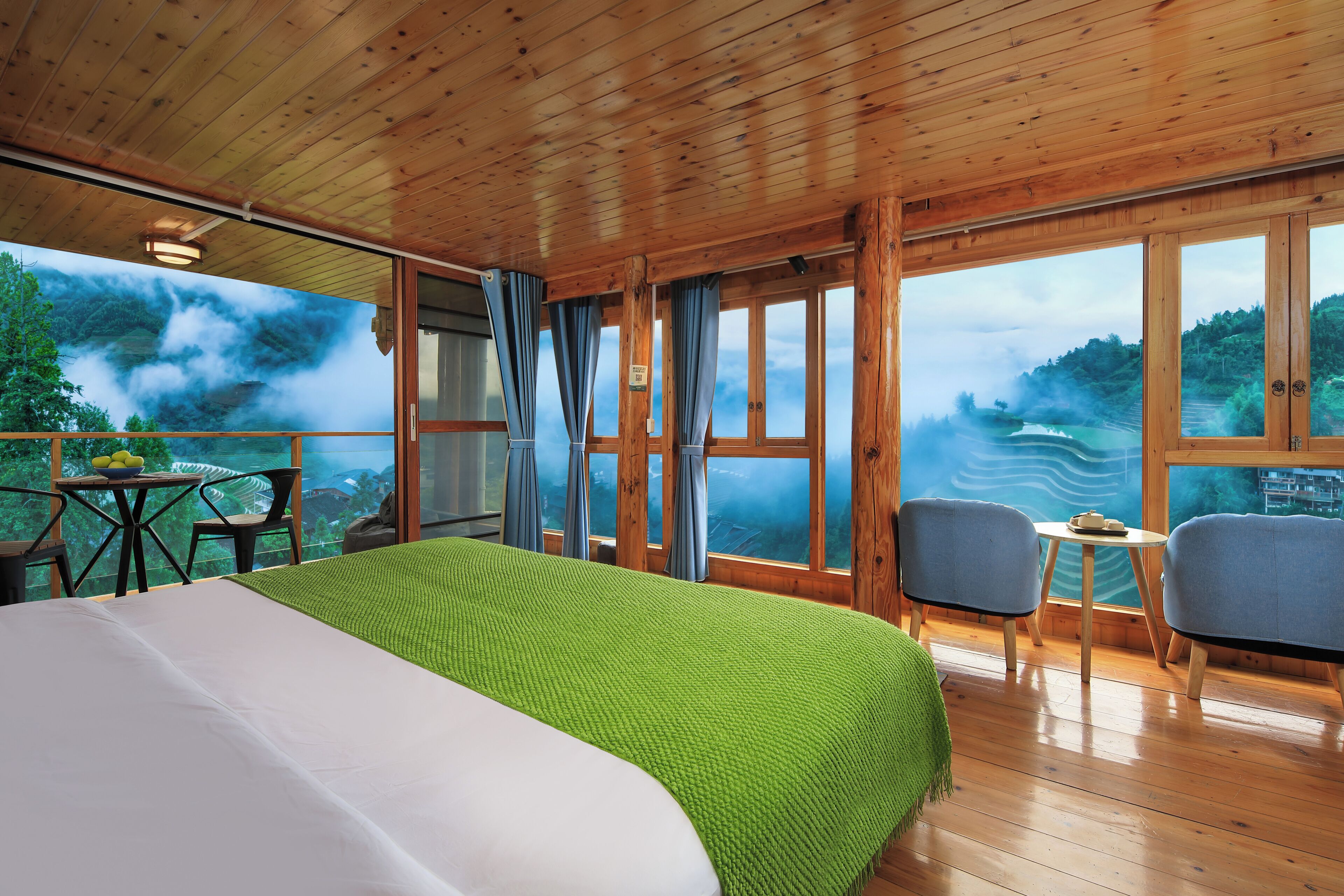 A wooden cabin room with a bed, two chairs, a small table, and large windows with a view of a forested landscape.