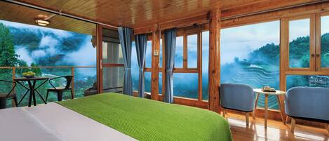 A wooden cabin room with a bed, two chairs, a small table, and large windows with a view of a forested landscape.