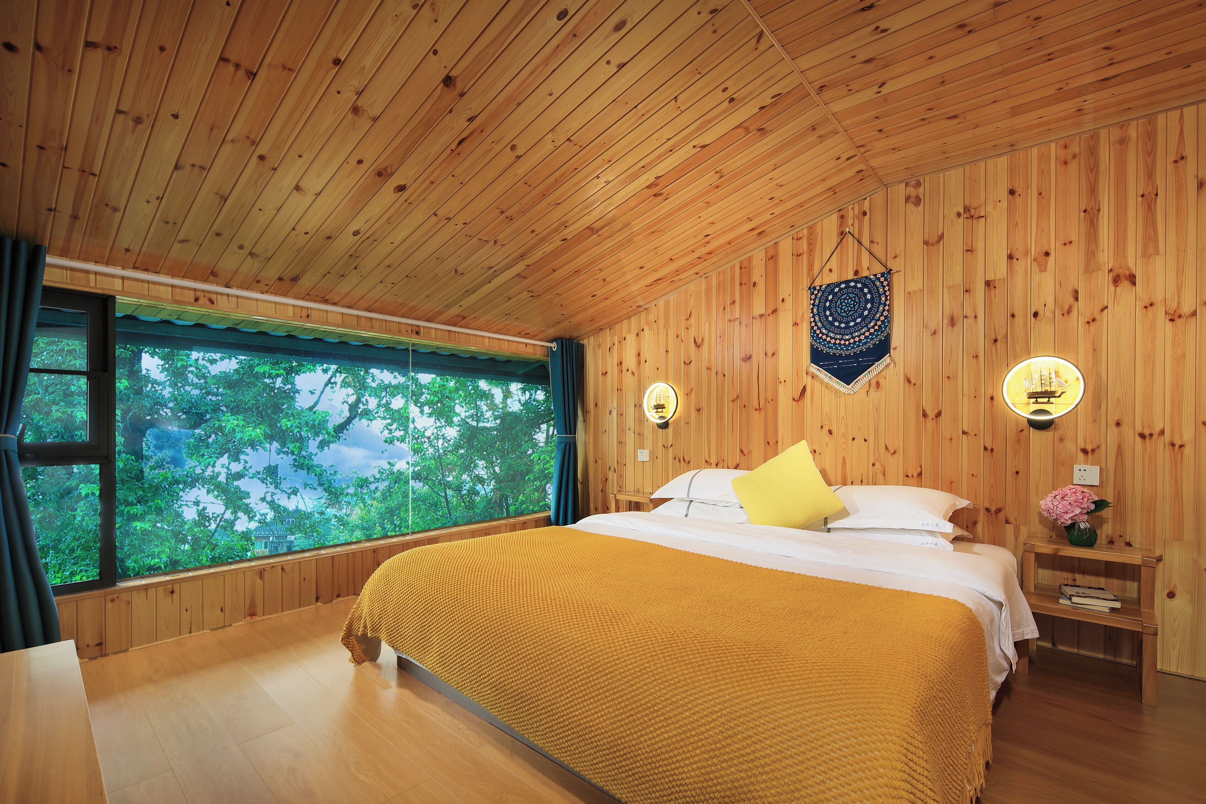 A wooden-paneled room with a bed, a window with a view of trees, a bedside table with a lamp, and a wall hanging.