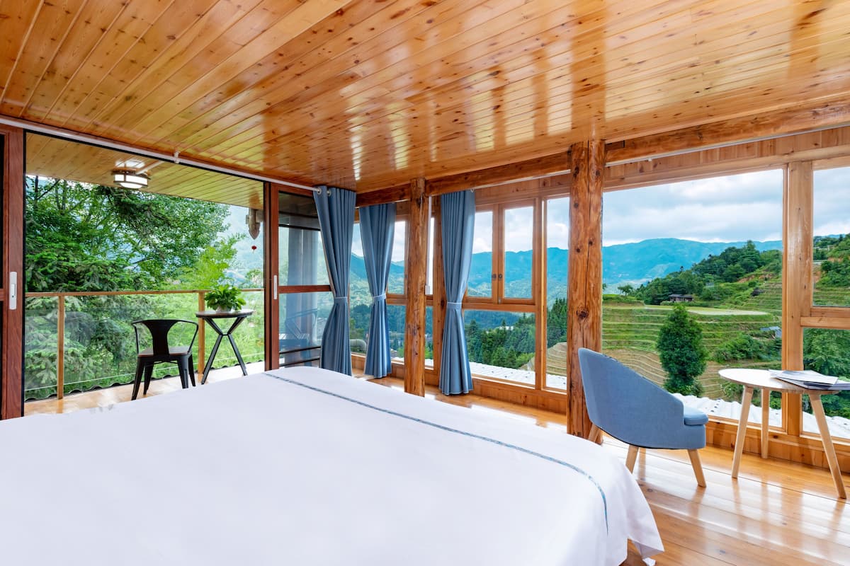 Una habitación de madera con una cama, una mesa y sillas, ventanas amplias con cortinas y vistas a colinas verdes.