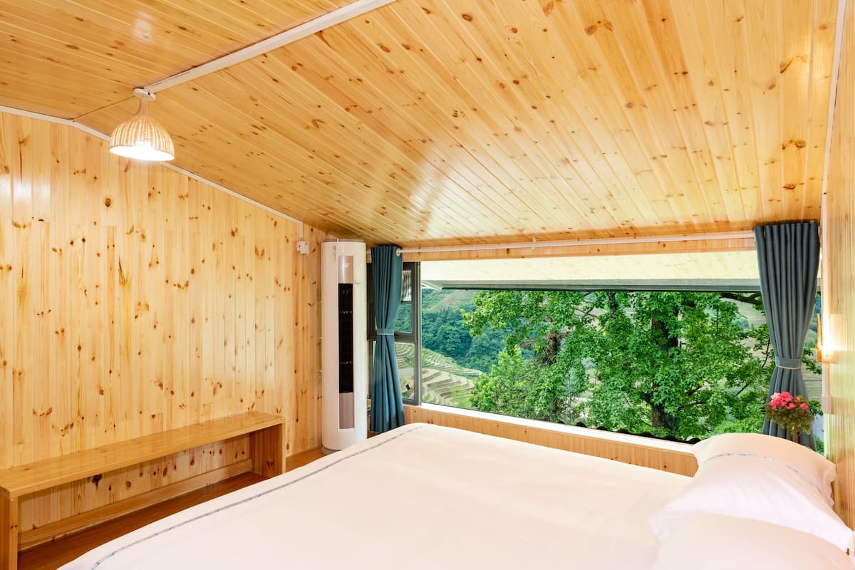 Una habitación de madera con una cama, un banco, una ventana con cortinas y una lámpara colgante.