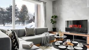 A modern living room with a grey sofa, a coffee table with pastries, a TV displaying 'NETFLIX,' and a view of a snowy landscape through large windows. - THE VELORA (Asahikawa)