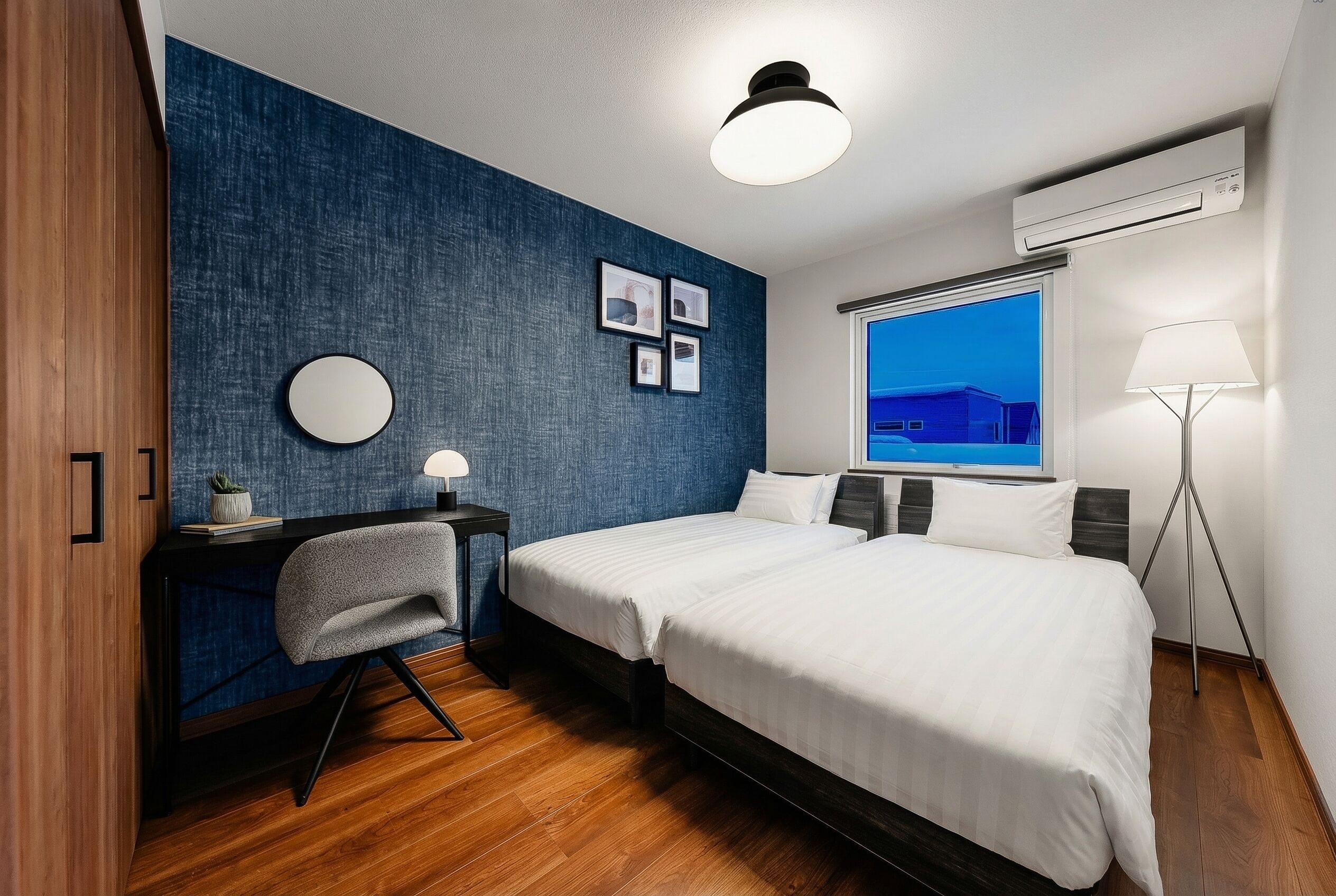 Et moderne hotelværelse med to senge, et skrivebord, en stol, et rundt spejl og et vindue med udsigt.