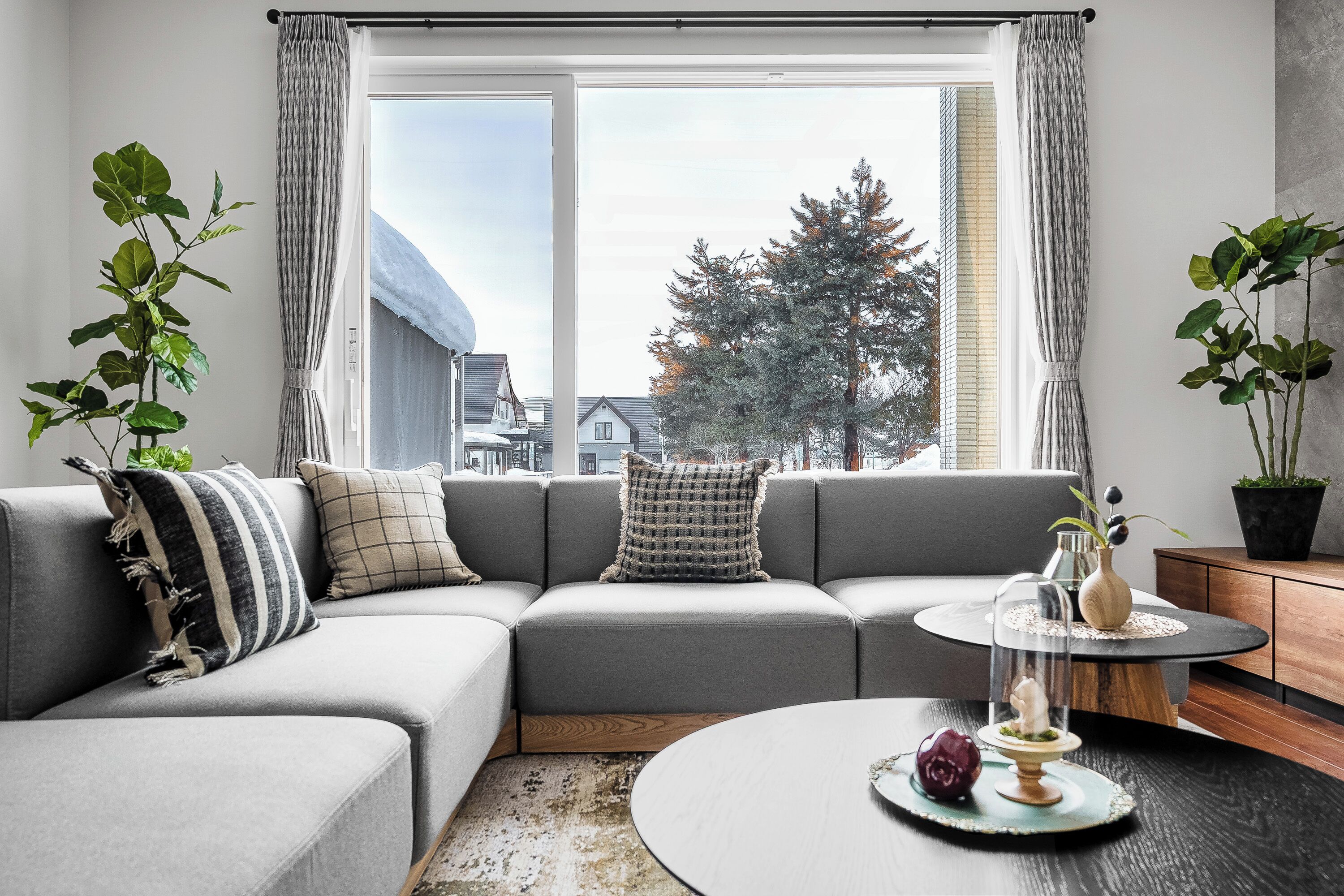 Un salon moderne avec un canapé d’angle gris, une table basse ronde et une vue sur un quartier résidentiel grâce à de grandes fenêtres.